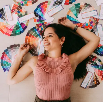 A woman with long, dark hair is lying on the floor, smiling and looking upwards. Surrounded by colorful fan-deck swatches spread out around her, she resembles a runway model displaying her talent. She wears a sleeveless pink ribbed top with a ruffled neckline.