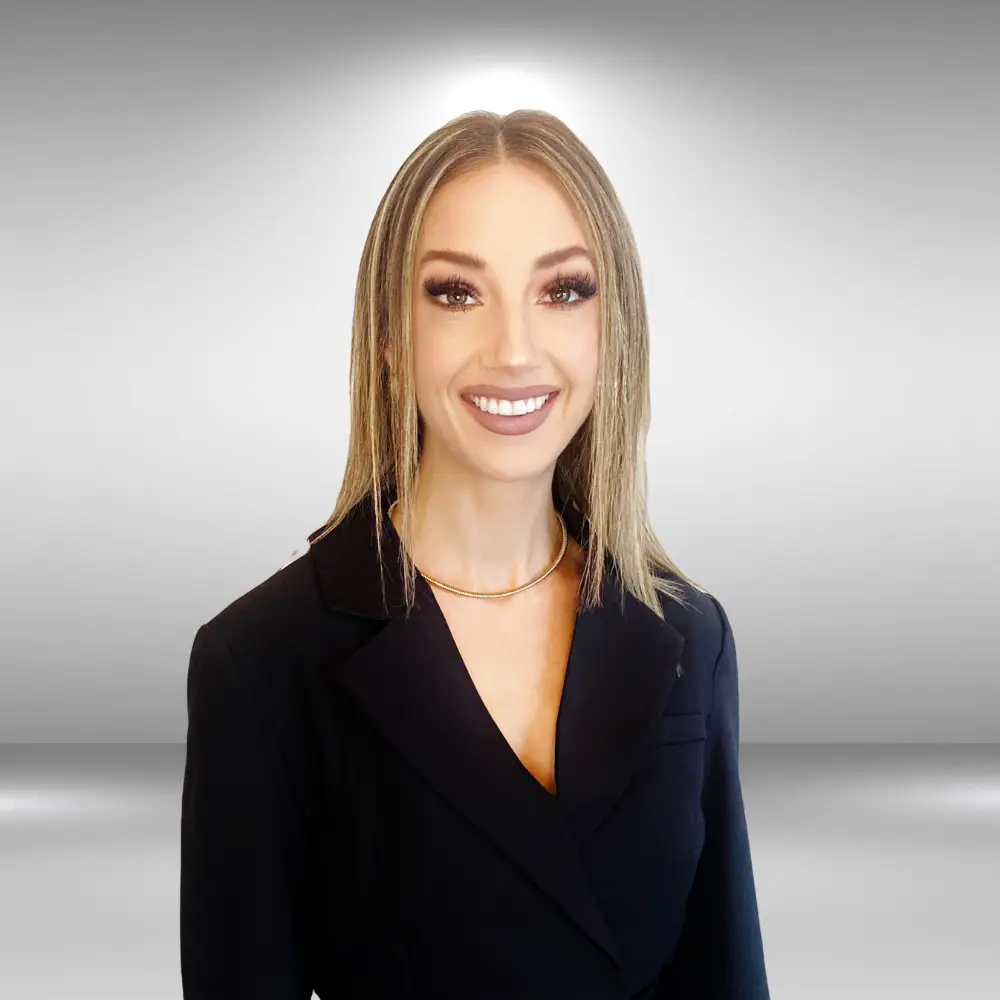 A smiling woman with straight, blonde hair, resembling a runway model, wears a black blazer and a gold necklace as she stands against a gradient gray background.