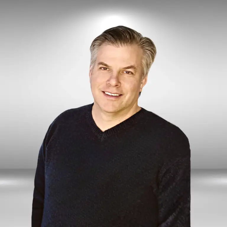 A man with short, gray hair and a friendly smile is wearing a dark sweater. He stands against a gradient gray background, exuding both professionalism and approachability. This talented actor captures everyone's attention with his poised demeanor.