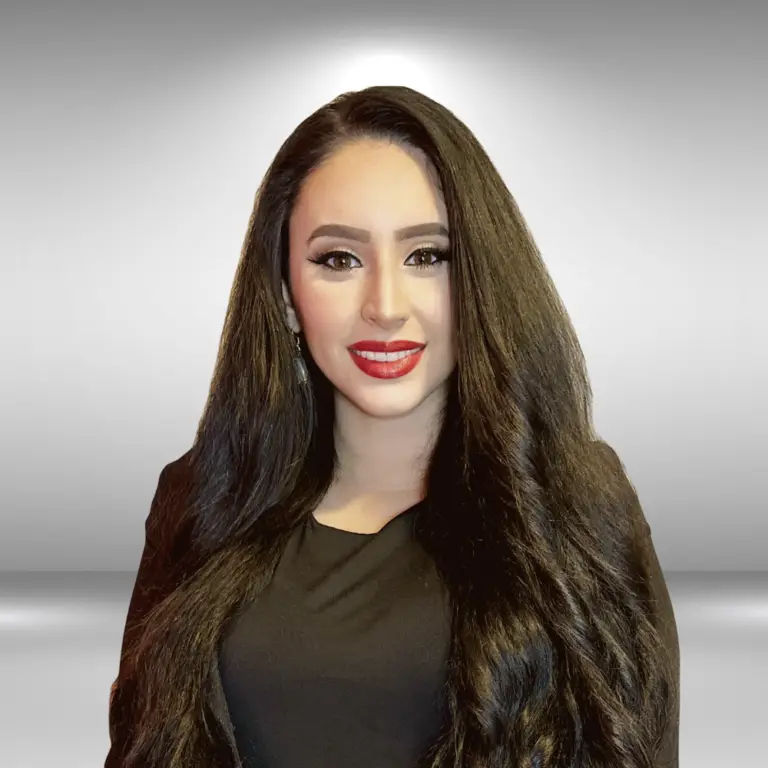 A woman with long, wavy black hair stands against a plain, light gray background. She is wearing a black top and has bright red lipstick, defined eyebrows, and eye makeup. Smiling confidently at the camera like an actor at an audition, she exudes charisma and presence.