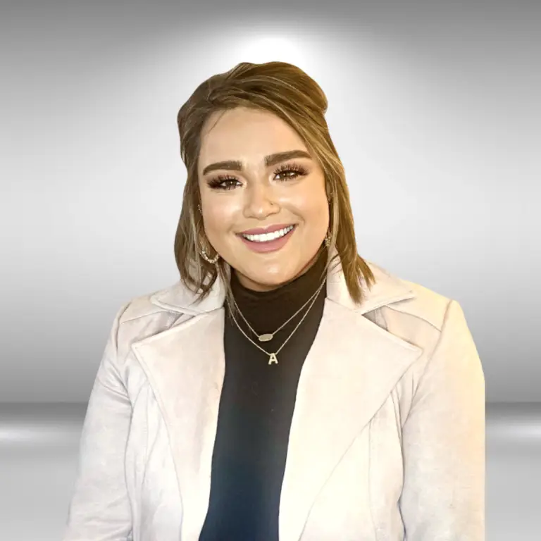 A young woman with brunette hair, styled in a half-up updo, is smiling warmly at the camera. She is wearing a light-colored jacket over a black top and layered necklaces, one of which has a pendant with the letter "A." She looks ready for an audition against the light, gradient gray background.