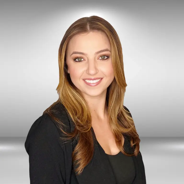 A woman with long, light brown hair and green eyes smiles at the camera. She is wearing a black top and a black blazer, exuding the poise of a runway model. The background is a simple gray gradient.