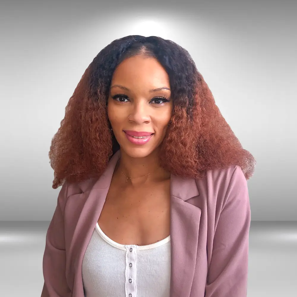 A woman with long, curly hair that transitions from black to reddish-brown smiles at the camera. She is wearing a white top underneath a light pink blazer, looking effortlessly chic like an influencer. The background is a gradient of light gray.