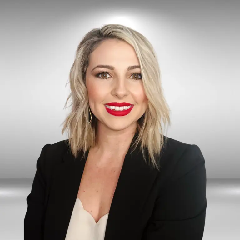 A talented woman with shoulder-length blonde hair, wearing a black blazer over a light-colored top, smiles confidently with bright red lipstick. She stands against a grey, gradient background.