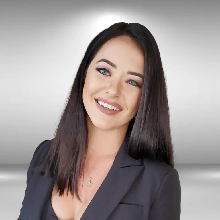 A smiling woman with long, straight dark hair is wearing a dark blazer over a black top. She has light-colored eyes and wears a delicate necklace. The background is a neutral gray gradient, perfect for an influencer's sleek and sophisticated image.