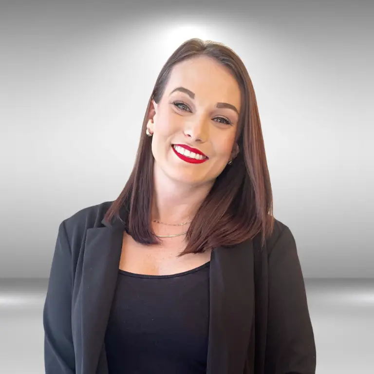 A smiling person with straight brown hair, red lipstick, and a black blazer over a dark top stands against a plain gray background, exuding the confidence and approachability of an actor ready for an audition.