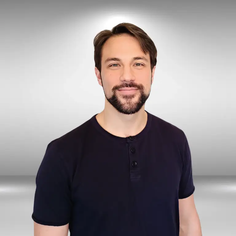 A man with short brown hair and a beard is smiling at the camera. He is wearing a navy blue short-sleeved shirt and standing against a plain grey background, looking ready for an audition as an actor or influencer.