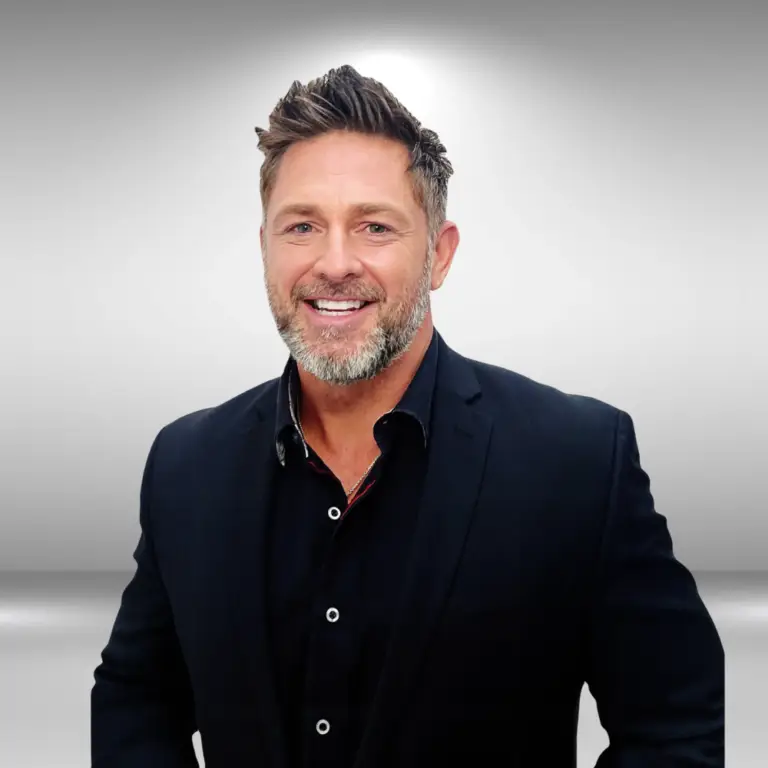 A man with short, styled hair and a beard smiles while wearing a black suit and shirt. He stands against a light gray, gradient background, looking every bit the poised actor ready for his next audition.