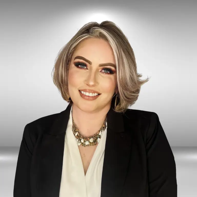 A woman with styled ash-blonde hair, wearing a black blazer over a white blouse, and a statement necklace. She has on makeup with a focus on bold eyes and is smiling. This stylish influencer stands against a simple gradient background from white to gray.