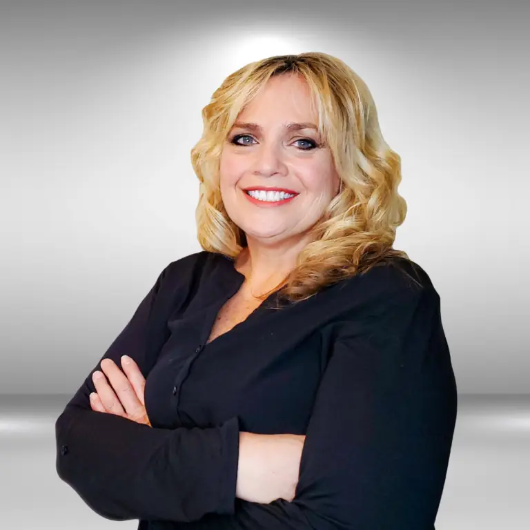 A talented woman with wavy blonde hair and blue eyes is smiling with arms crossed. She is wearing a black long-sleeved top and is standing against a light gray gradient background, exuding the confidence of an influencer.