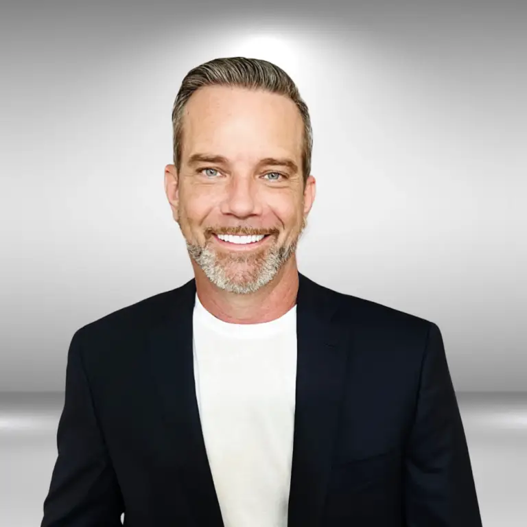 A man with short brown and gray hair and a trimmed beard, wearing a black blazer over a white shirt, stands smiling in front of a gradient gray background. He exudes confidence, ready for his audition as an aspiring model influencer.