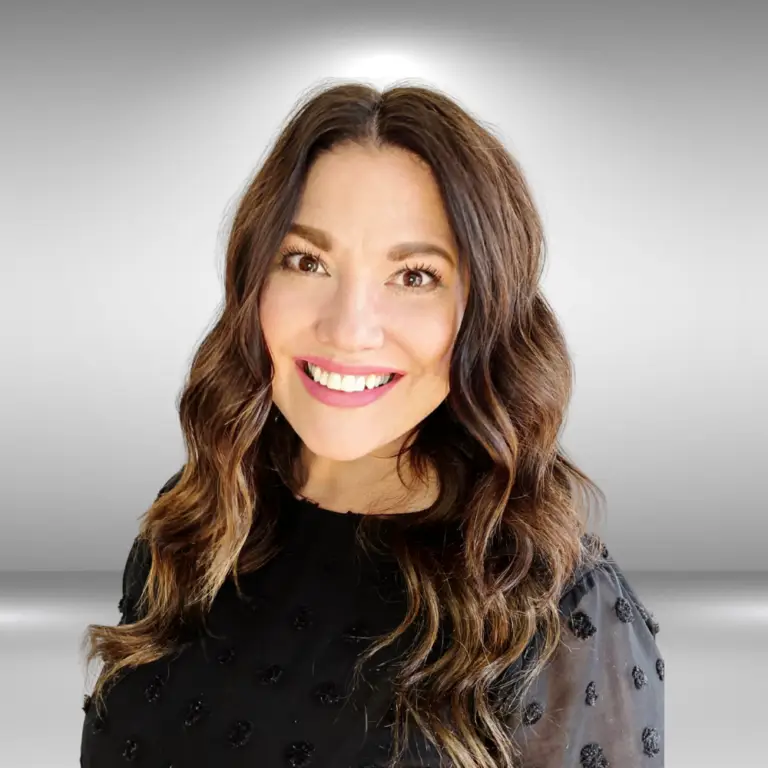 A talented woman with long wavy brown hair smiles at the camera. She is wearing a black blouse with a subtle textured pattern. The background is a gradient of light grey to white, creating a soft halo effect around her head, as if she's ready for an audition.