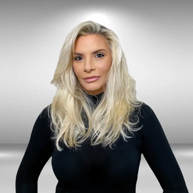 A model with long, wavy, platinum blonde hair stands against a plain, light gray background. She is wearing a black, long-sleeved top and looking directly at the camera with a neutral expression, as if ready for an audition.