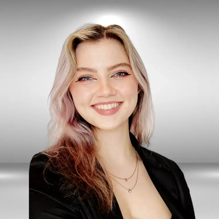 A young woman with long, wavy blonde hair and light purple undertones smiles warmly. She wears a black blazer and a delicate necklace with small pendants. The background is a gradient of light to dark gray, reminiscent of the polished atmosphere before a talent audition.