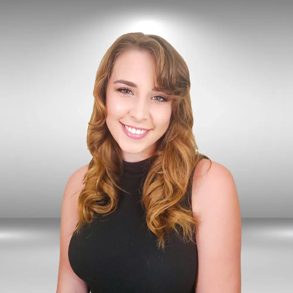 A young woman with long, wavy, light brown hair and fair skin smiles at the camera. Wearing a sleeveless black top, she exudes confidence as if she's ready for a runway audition, standing against a plain, gray background.