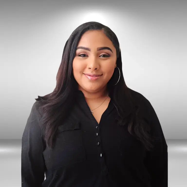 A talented woman with long, dark hair and hoop earrings stands against a gradient grey background. She is wearing a black button-up shirt and smiles gently at the camera, exuding the poise of an actor ready for the runway.