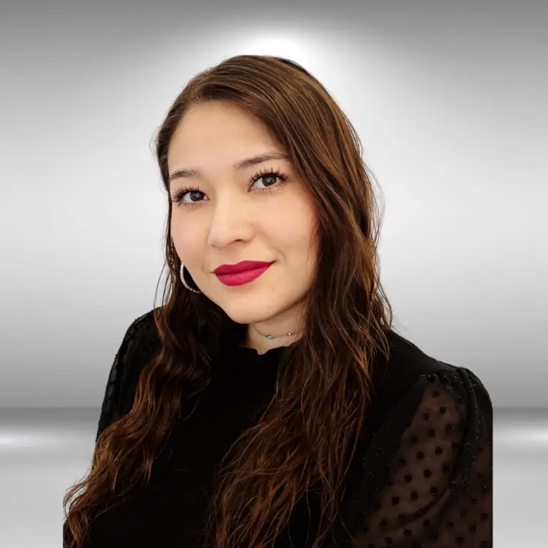 A woman with long, wavy brown hair and wearing a black top with sheer sleeves, smiling against a plain, light gray backdrop. She has hoop earrings and is wearing red lipstick — ready for her audition as an actor.