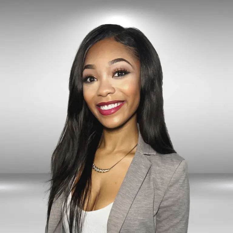 A woman with long, straight dark hair smiles warmly at the camera. She is wearing a light gray blazer over a white top, and a delicate necklace. The background is a simple gradient of gray to white, giving a professional appearance that makes her look ready for an audition.