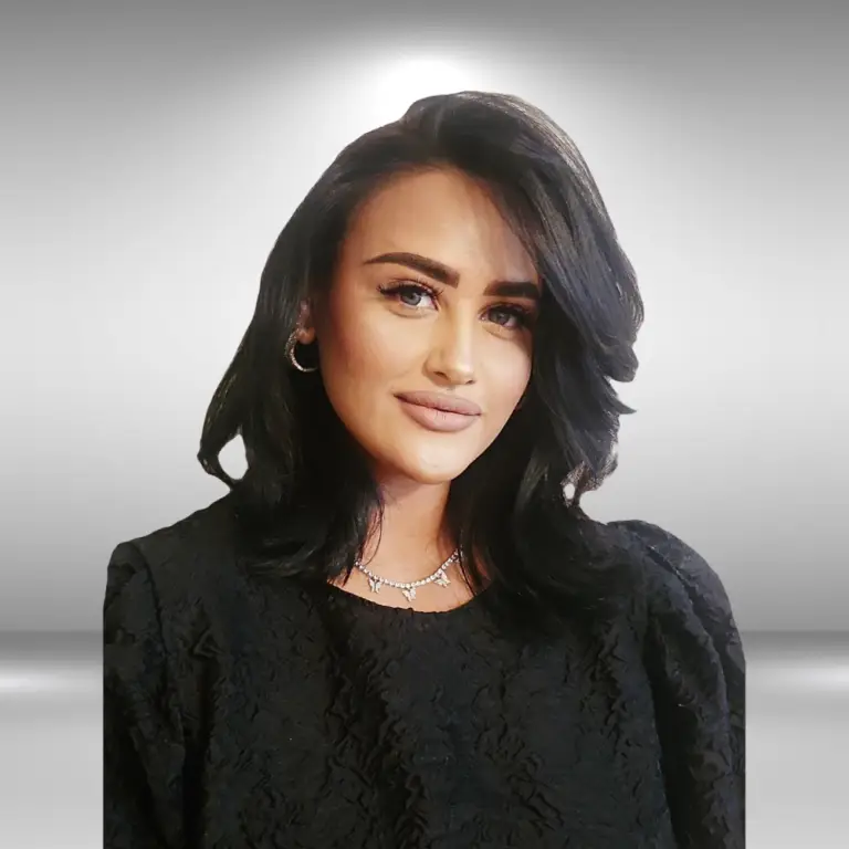 A woman with long, dark hair styled in waves is posing for the photo. She is wearing a black, textured top with a round neckline and a silver necklace. The background is a smooth gradient from dark to light gray, highlighting her against the backdrop—perfectly capturing her talent for an upcoming audition.