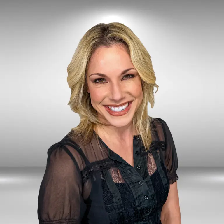 A smiling model with blonde hair is wearing a black, sheer blouse. She poses against a gradient grey background.