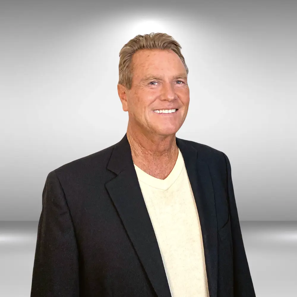 A smiling man with short, light brown hair is wearing a black blazer over a light yellow shirt. He stands confidently against a simple, light grey gradient background, ready for his audition as an influencer.