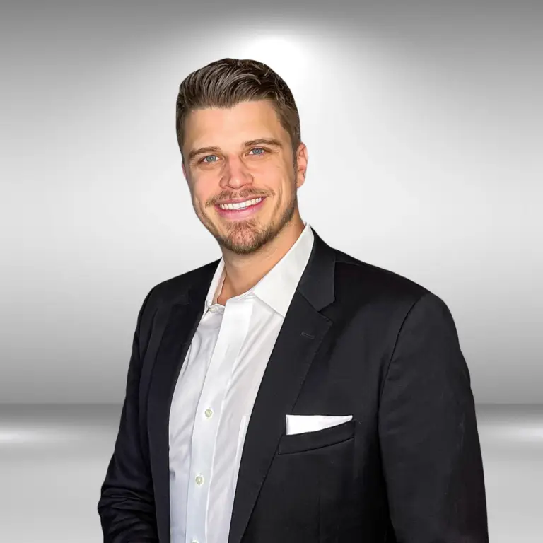 A talented man with short hair, a beard, and a mustache, dressed in a black suit jacket, white dress shirt, and pocket square, smiles at the camera. The background is a plain, gradient gray.