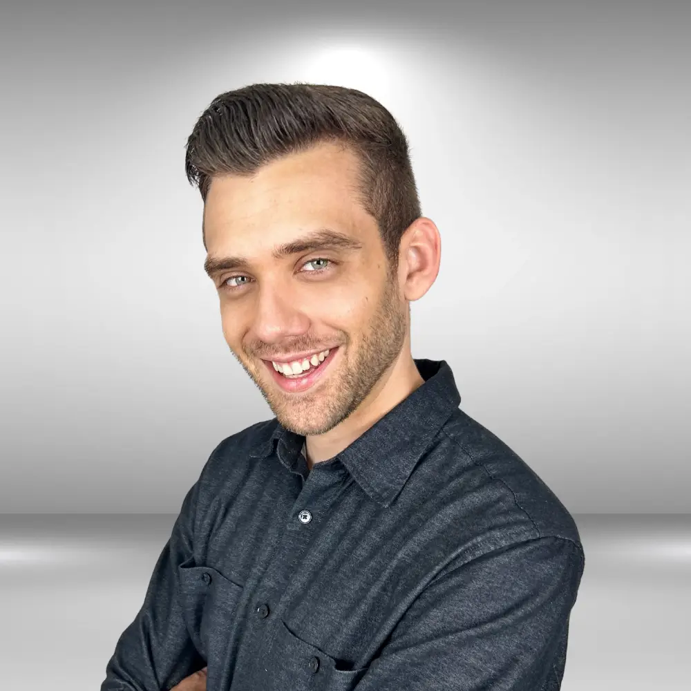 A man with short dark hair and a beard smiles while crossing his arms. He is wearing a dark button-up shirt and stands in front of a plain grey background, ready to showcase his talent for the audition.