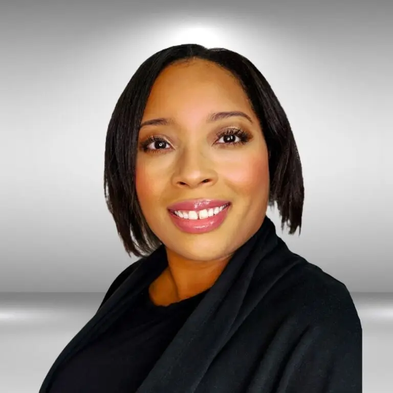 A person with shoulder-length dark hair is smiling and looking at the camera. They are wearing a black top with a black draped fabric over their shoulders, giving off an air of talent. The background is a plain, light-colored gradient.