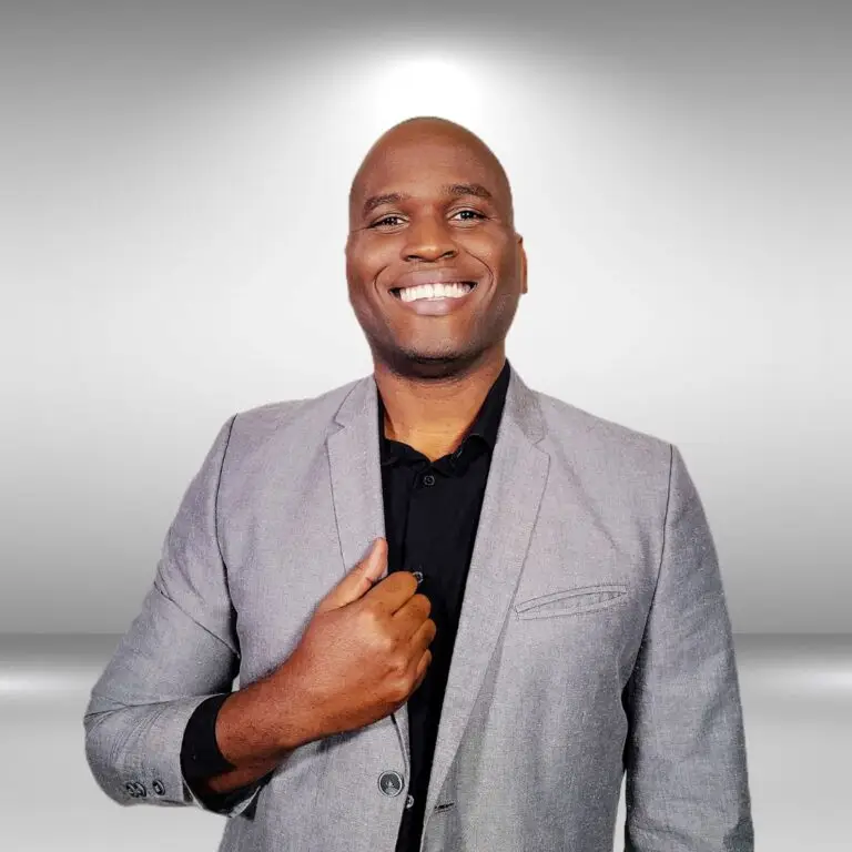 A smiling actor stands confidently in front of a plain, light gray background. They are wearing a light gray blazer over a black shirt and have their right hand resting on their chest.