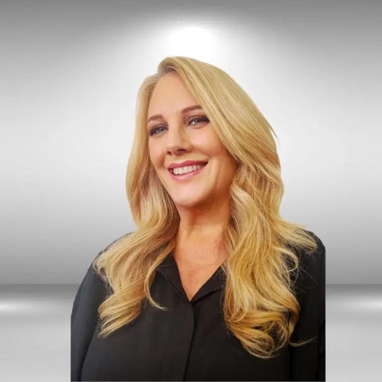 A woman with long, wavy blonde hair smiling at the camera. She is wearing a black shirt, and the background is a gradient of light grey, giving her the appearance of a model ready for an audition.