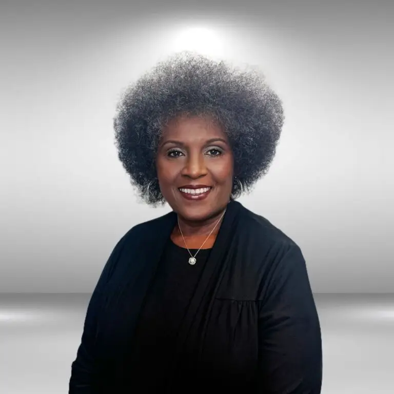 A woman with short, curly hair smiles at the camera. She wears a black top with a matching black cardigan and a small pendant necklace. The influencer stands confidently as if ready for the runway, against a background that transitions from light gray to white.