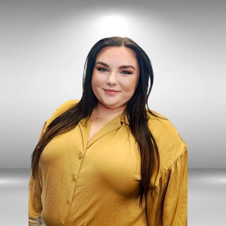 A woman with long, dark hair, wearing a mustard yellow blouse, stands smiling against a neutral, gradient background, exuding the elegance of a runway model.