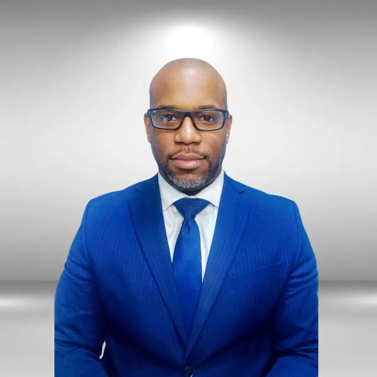 A man with a shaved head and beard, wearing glasses and a blue suit with a matching tie, poses confidently against a plain grey background. With the poise of a runway model, he looks directly at the camera with a serious expression.