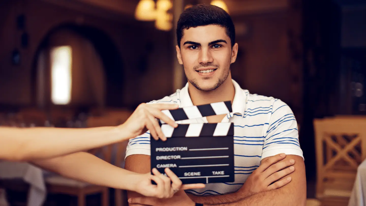 A man in a white and blue striped shirt is sitting in a dimly lit room, smiling with arms crossed. A hand holds a clapperboard in front of him, indicating he is likely auditioning on a film set or participating in a video recording.