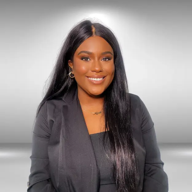 A woman with long, straight black hair is smiling at the camera. She is wearing a black blazer over a black top and has gold hoop earrings and a delicate necklace. The background is a neutral gray gradient, highlighting her poised look reminiscent of an influencer auditioning for her next big role.