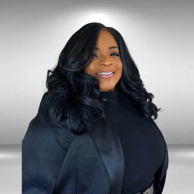 A talented individual with long, wavy black hair smiles while wearing a black blazer and shirt. They stand against a grey background with a subtle spotlight effect behind them.