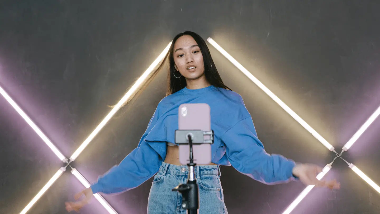 A model wearing a blue cropped sweater and jeans stands in front of a smartphone mounted on a tripod, gesturing while recording. The background features intersecting neon light bars arranged in a diamond pattern against a dark wall.