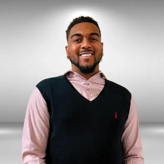 A man with short curly hair and a beard smiles at the camera. Dressed in a light pink button-up shirt under a black V-neck sweater, he exudes confidence. The background is a grey, gradient studio setting, perfect for an influencer's profile or model audition portfolio.