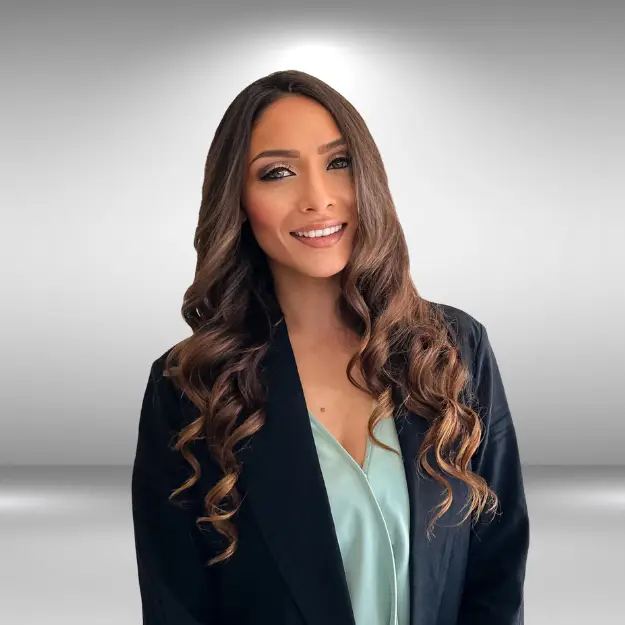 A professionally dressed individual with long, wavy brown hair smiles warmly at the camera. They are ready for their audition, wearing a light blue top under a black blazer, set against a neutral grey gradient background.