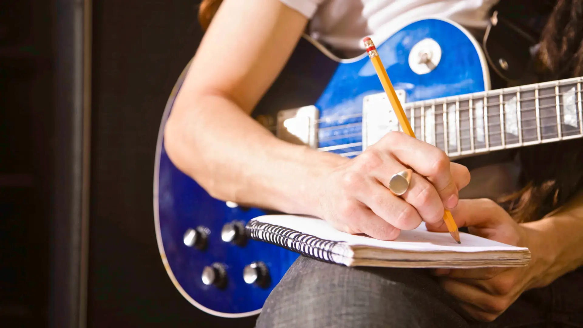 A talented individual is sitting with a blue electric guitar, meticulously writing in a notebook with a pencil. Dressed in jeans and a white t-shirt, and sporting a ring on one finger, the focus is on their hands, the guitar, and the notebook.