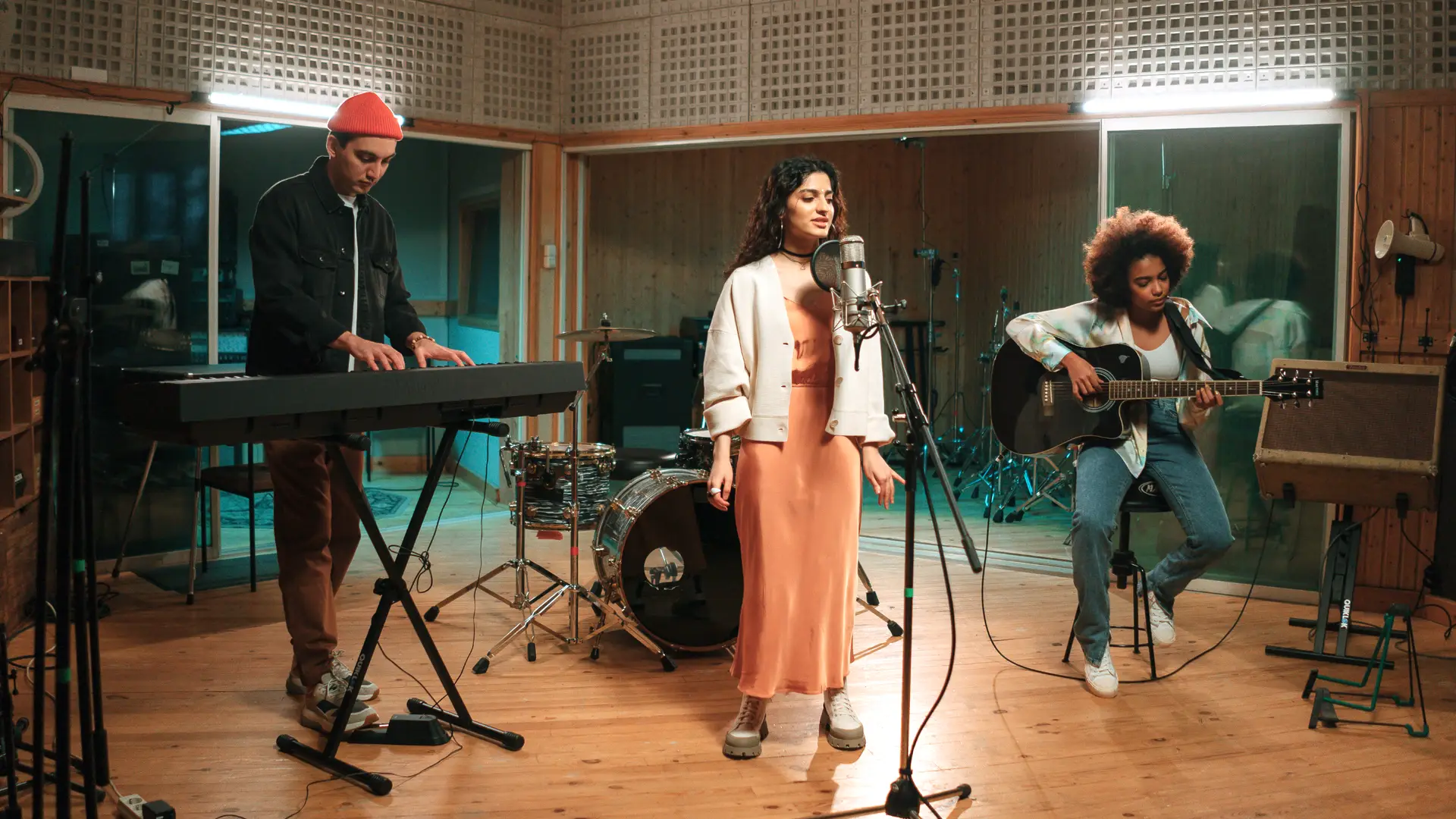 A band showcases their talent in a recording studio. A woman sings into a microphone at the center, a man in a red beanie plays a keyboard to the left, and another woman with an afro hairstyle models her musical prowess as she strums an acoustic guitar while seated to the right. Drums are visible in the background.